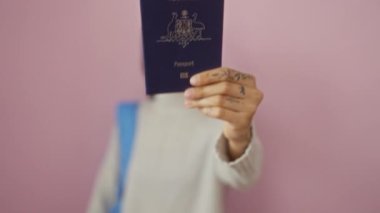 Avustralyalı pasaportu olan genç bir kadın, izole edilmiş pembe arka plana gülümsüyor, gri kazak ve mavi sırt çantası giyiyor, odak noktasında dövmeli elini gösteriyor.