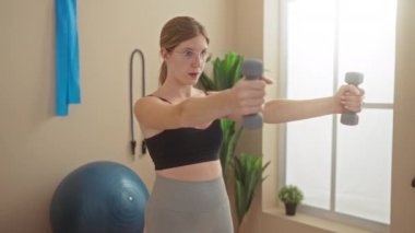 Spor salonunda dambıllarla egzersiz yapan bir kadın, spor kıyafetleri içinde sarışın, genç, çekici bir kadın, modern spor merkezinde spor salonuna odaklanmış, ekipmanlar odada görünür.