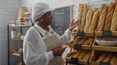 Bir fırında kadın fırıncı, arkasında ekmek rafları ile ekmek çeşitlerini açıklarken 