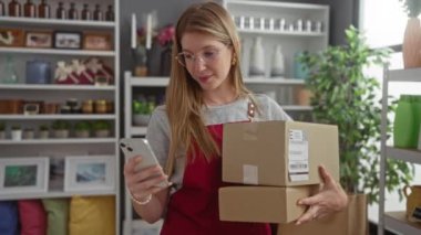 Gözlük ve önlük takan genç bir kadın elinde karton kutular ve arka planda dekoratif eşya rafları olan bir ev dekorasyon mağazasında akıllı telefon kullanıyor.