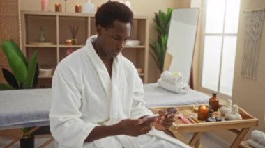 Kapalı bir güzellik merkezinde sağlık araçlarıyla çevrili akıllı telefon kullanan spa elbiseli genç bir adam.