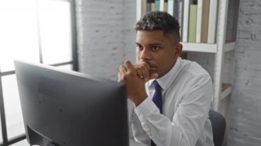 Ofisteki genç adam düşünceli bir şekilde bilgisayar üzerinde çalışıyor, beyaz gömlek ve mavi kravat takıyor, arka planda kitap rafları var, profesyonel ve odaklanmış çalışma ortamı sergiliyor.