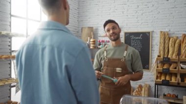 Baker, sergilenen raflarda çeşitli ekmek ve hamur işleriyle dolu parlak bir fırında müşteriyle konuşuyor..