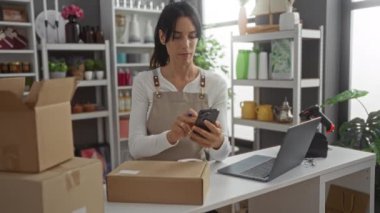 Kadın etiketleri, arka planda renkli eşyalar bulunan, beyaz bir üst ve bej önlük giyen, ev dekorasyon mağazasında telefon ve kalem kullanan paketlerdir.