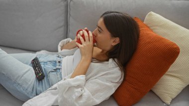 Evdeki rahat bir kanepede rahat rahat kahve içen genç bir kadın. Yastıklı ve uzaktan kumandalı rahat bir oturma odasında.