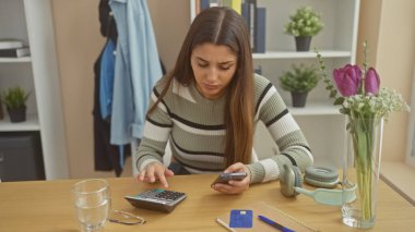 İspanyol kadın evde akıllı telefondan mali durumları hesaplıyor, etrafı kişisel eşyalarla çevrili, günlük ev yaşamını resmediyor..