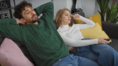 Modern oturma odalarında rahat bir çift, kadın kumandayı tutuyor ve adam rahatça esniyor..