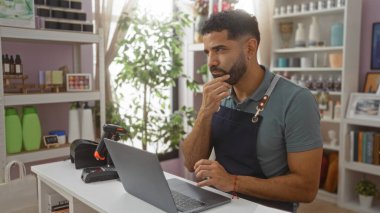 Genç adam bir ev dekorasyonu dükkanında, ürünlerle çevrili, elini çenesinin altına sararken bir dizüstü bilgisayarla çalışıyor..