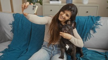 Genç bir İspanyol kadın, Fransız bulldogunu sıcak bir oturma odasında tutarken selfie çekiyor..