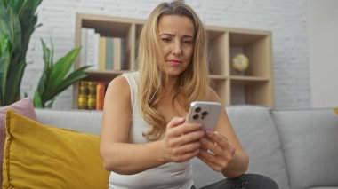 Odaklanmış beyaz bir kadın, rahat bir oturma odasında akıllı telefonunu kullanarak koltuğa oturmuş..