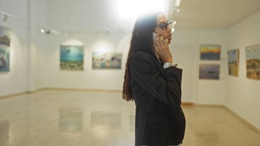 Bir sanat galerisinde telefonla konuşan, duvarlarında resimler olan, müzenin veya stüdyonun içini gösteren genç bir kadın..