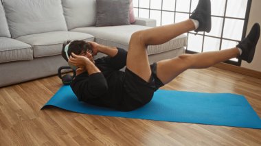 Genç, İspanyol bir adam oturma odasında mavi bir paspasın üzerinde kas egzersizi yapıyor. Sağlıklı bir yaşam tarzını ve rahat bir ev ortamını ön plana çıkarıyor..