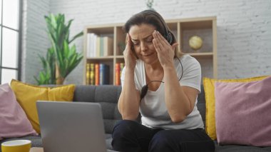 Evde kulaklıklı stresli bir kadın. Arka planda bulanık kitaplık olan tapınaklara dokunuyor..