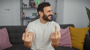 Gülümseyen sakallı adam, orta parmaklarıyla oturma odasında el kol hareketi yapıyor, rahat ve oyuncu davranışlar sergiliyor..