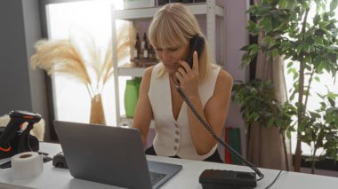 Kadın telefonda konuşuyor ve arka planda bitkiler ve dekoratif malzemelerle çevrili bir ev dekoratörü dükkanında dizüstü bilgisayarla çalışıyor..