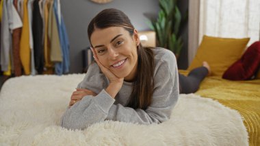 Esmer saçlı, gri kazak giyen, rahat bir yatak odasında yatağında uzanan güzel, genç bir İspanyol kadın..