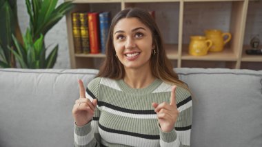 Genç İspanyol kadın, kapalı bir koltukta otururken yukarı doğru işaret ediyor. Mutlu bir ifade ve günlük kıyafetle..