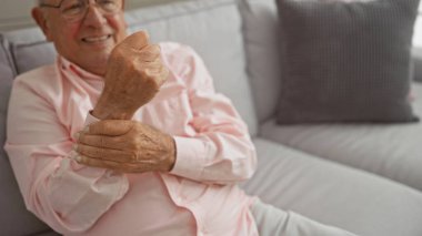 Gözlüklü ve pembe gömlekli yaşlı beyaz bir adam rahat bir kanepede oturuyor. Parlak bir oturma odasında, rahat bir tavırla gülümsüyor..