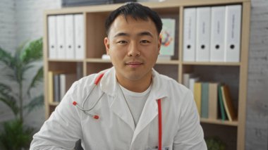 Klinikteki yakışıklı genç bir Çinli adamın portresi, arka planda organize dosyalar ve bitkilerle birlikte Asyalı bir doktoru çalışma odasında sergiliyor..
