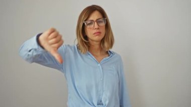 Genç bir kadın, sarışın, gözlük takıyor, baş parmağı aşağıda, negatif jest yapıyor, mutsuz, kızgın yüz ifadesi, beyaz izole edilmiş arka plana bakıyor, kötü bir işaret gösteriyor.