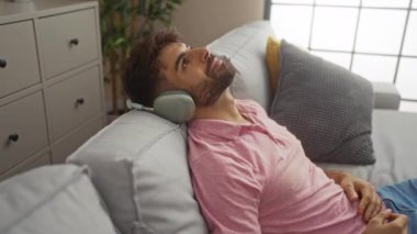 Genç adam evde dinleniyor, rahat bir oturma odasında kanepede uzanmış kulaklıkla müzik dinliyor.