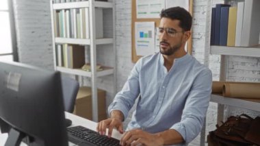 Genç adam modern ofis ortamında stresli görünen gözlükleri kaldırıyor, kitap rafları, grafikler ve bir bilgisayar ile çevrili, yoğun bir çalışma ortamını yansıtıyor..
