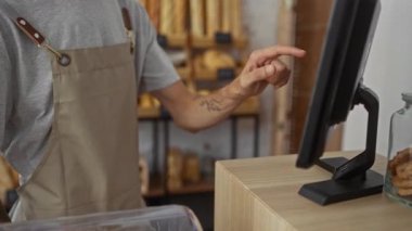 Fırında çalışan genç bir adam dokunmatik ekran kaydına dokunuyor, gri bir gömlek ve önlük giyiyor, arka planda ekmek somunlarıyla dolu ahşap raflar var..