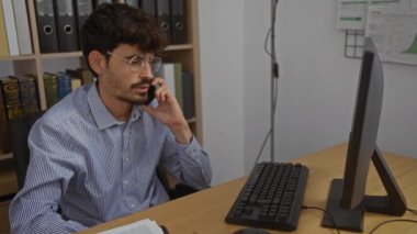 Ofisteki sakallı genç adam içeride bilgisayarın arka planında kitaplarla telefonda konuşuyor.