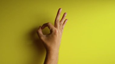 Parlak sarı bir arkaplana karşı el hareketi yaparak parmakları açık ve minimalist bir şekilde vurguluyor..