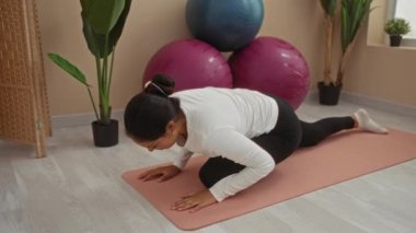 Spor salonundaki bir paspasın üzerinde yoga yapan genç bir kadın. Toplar ve bitkilerle çevrili, beyaz bluzlu ve siyah taytlı.