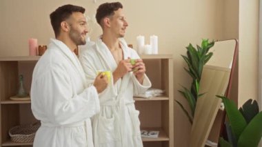 Bornoz giyen erkekler spa odasında çay içiyorlar. Mumlar, bitkiler ve raflar.