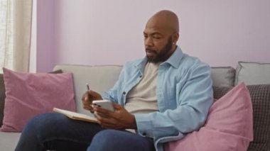 Oturma odasının iç kısmında akıllı telefon kullanırken not defterine yazan yakışıklı bir adam.