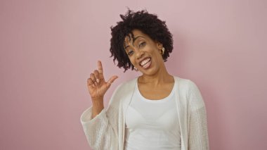 Kıvırcık saçlı, güzel bir Afro-Amerikalı kadın pembe bir arka plana karşı poz veriyor, özgüvenle gülümsüyor..