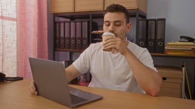 Sakallı genç bir İspanyol adam organize bir ofiste dizüstü bilgisayarla çalışırken kahve içiyor. Dosyaları ve renkli perdeleri var..