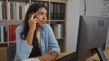 Ofiste akıllı telefondan konuşan, arka planda kitap rafları olan, gülümseyen ve bilgisayar masasında çalışan, profesyonel ve sıradan bir çalışma ortamı sergileyen bir kadın.