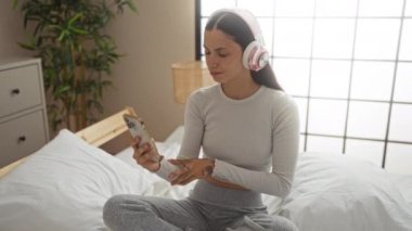 Evde yatak odasında telefonla müzik dinleyen genç bir kadın.