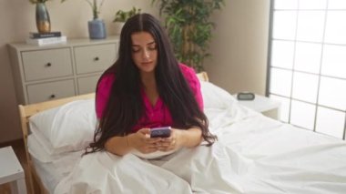 Beyaz yatak odası ve saksı bitkileriyle rahat bir yatak odasında akıllı telefon kullanan bir kadın, modern iç dekorda bir sabah sahnesini yakalıyor.