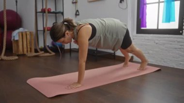 Spor salonundaki yoga minderinde egzersiz yapan bir kadın arka planda fitness ekipmanlarıyla antrenman rutinini gösteriyor.