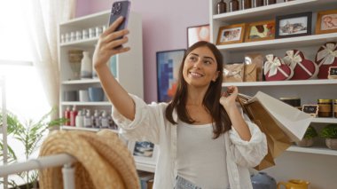 Elinde alışveriş poşetleri olan, kalp şeklindeki kutular, çerçevelenmiş sanat eserleri ve saksı bitkileri gibi güzel dekoratif eşyalarla süslenmiş bir mağazada selfie çeken bir kadın.