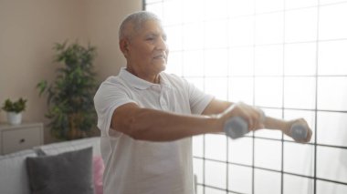 İspanyol, gri saçlı, yakışıklı orta yaşlı bir adam. Parlak bir oturma odasında halterlerle egzersiz yapıyor..