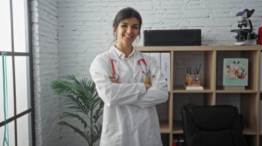 A young hispanic female doctor with brunette hair stands confidently with crossed arms in a modern clinic office, showcasing a welcoming and professional atmosphere. clipart