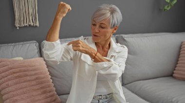 Yaşlı bir kadın oturma odasında kolunu esnetiyor, güç ve kendine güven gösteriyor, gri ve pembe bir dekor, kısa gri saçlar ve rahat bir apartman dairesinde beyaz gömlek ile bir kanepeye oturuyor.