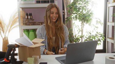 Ev dekorasyonu mağazasındaki genç sarışın kadın dizüstü bilgisayarında çalışırken gülümsüyor. Dekoratif eşyalar ve bitkilerle dolu. Üretkenlik ve hoş iç dekorasyon karışımı sergiliyor..
