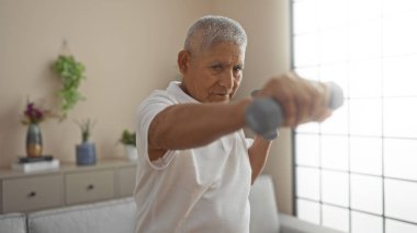 Beyaz saçlı İspanyol bir adam, parlak bir oturma odasında dambıllarıyla egzersiz yapıyor, evde sağlıklı aktivitelerle meşgul yetişkin birini gösteriyor..