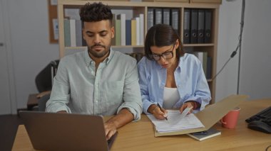 Bir ofis masasında çalışan bir adam ve bir kadın bir dizüstü bilgisayar ve bir çalışma ortamında belgeler ile birlikte çalışıyorlar.