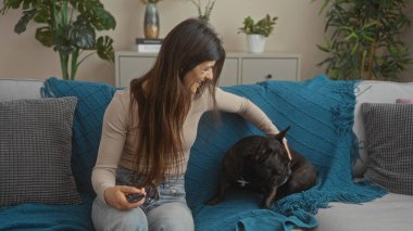 Genç bir İspanyol kadın oturma odasında elinde uzaktan kumandayla oturup buldogunu okşuyor..