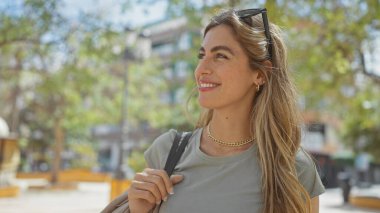 Gülümseyen genç bir kadın yeşilliklerle çevrili bir şehir parkında güneşli bir günün tadını çıkarıyor.