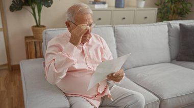 Yaşlı, kır saçlı, beyaz, oturma odasında oturan bir adam, koltukta oturmuş bir mektup okuyor.