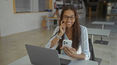 Kapalı bir ofiste telefonuna bakarken gülümseyen gözlüklü genç bir kadın.