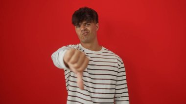 Çizgili tişört giyen genç, İspanyol bir adam, izole edilmiş kırmızı arka plana karşı baş parmak işareti yapıyor..